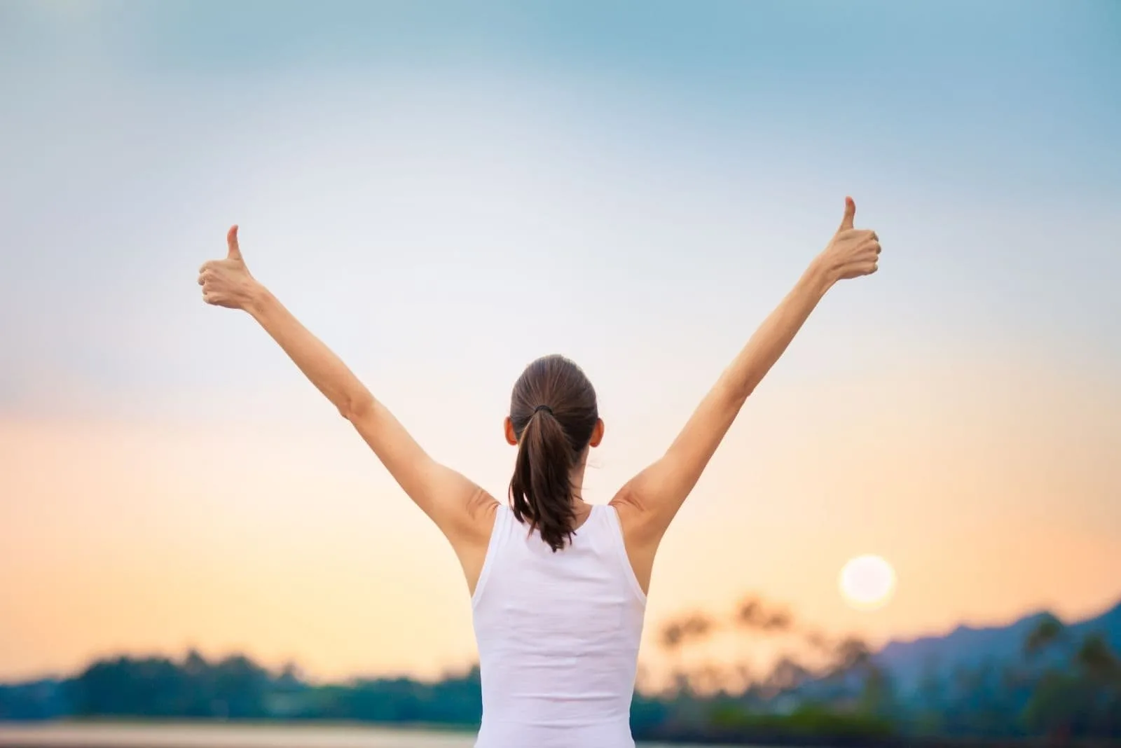 athletic woman raising her hands with thumbs up faciing the sunrise/sunset outdoors