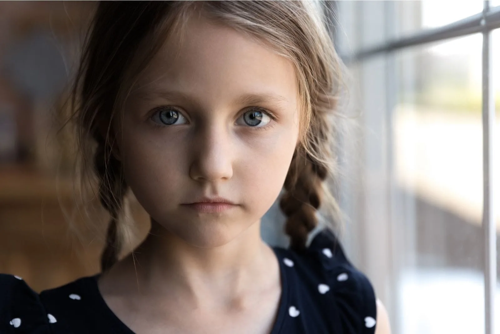 cropped image of a sad child looking at the camera standing beside the windows