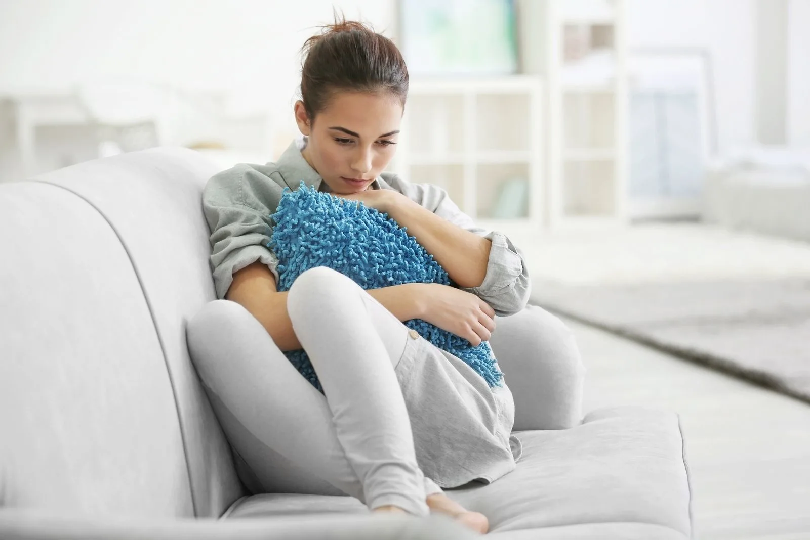 depressed young woman sitting with feet tucked in the gray couch embracing pillow in the living room
