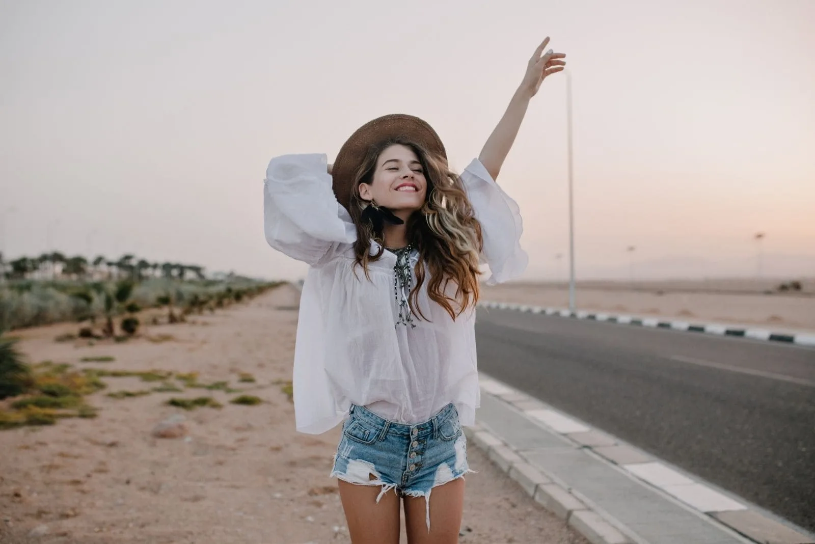 happy woman stands near the street along the highways holding her hat and raising one arm