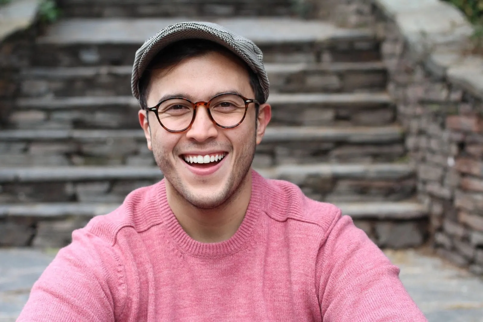 hip man smiling wearing hat and eyeglasses sitting near the cement stairs