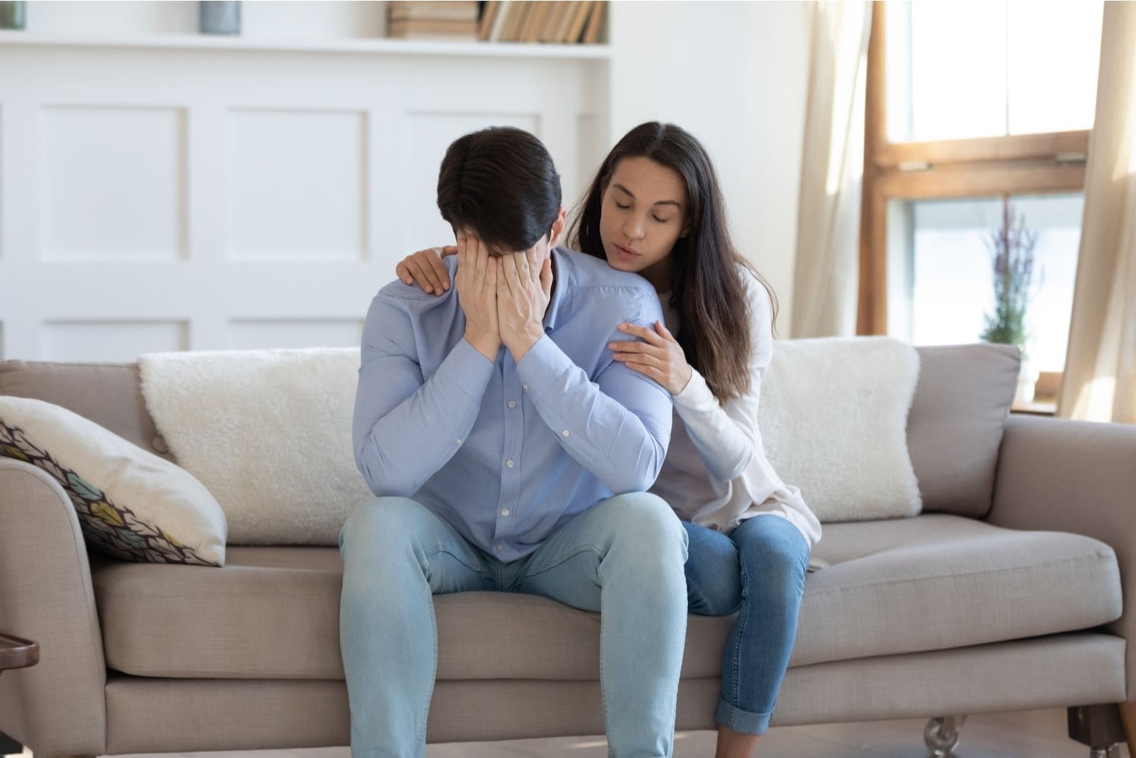 loving young wife hugging her depressed sad husband sitting inside the living room 