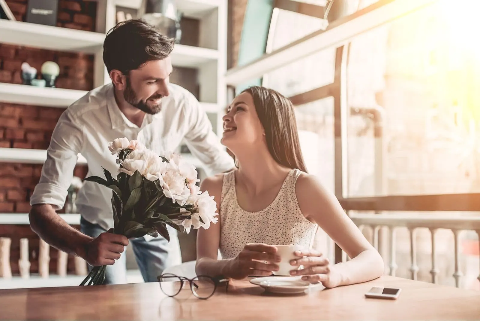 man giving flower to a beautiful woman sitting inside the cafe lloking surprised