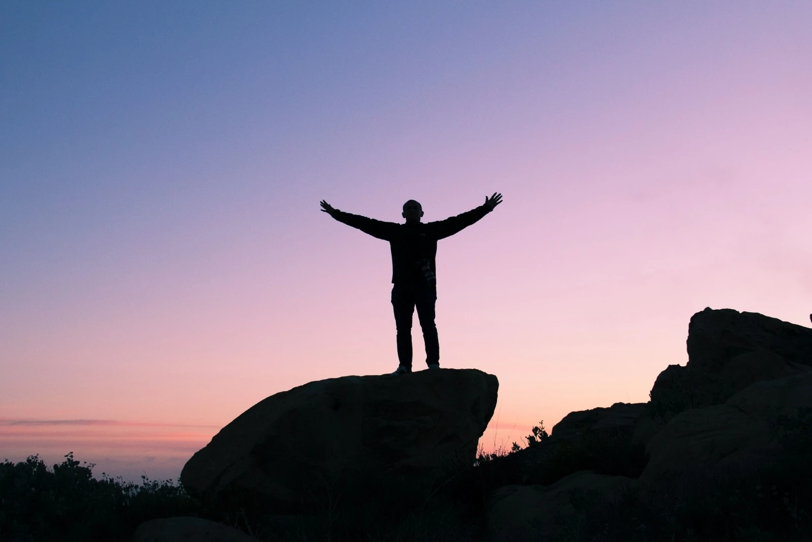 man raising his hands while standing on rock during sunset