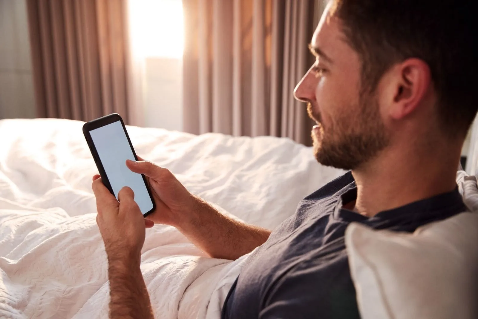 man sitting on bed with his cellphone on sideview inside bedroom