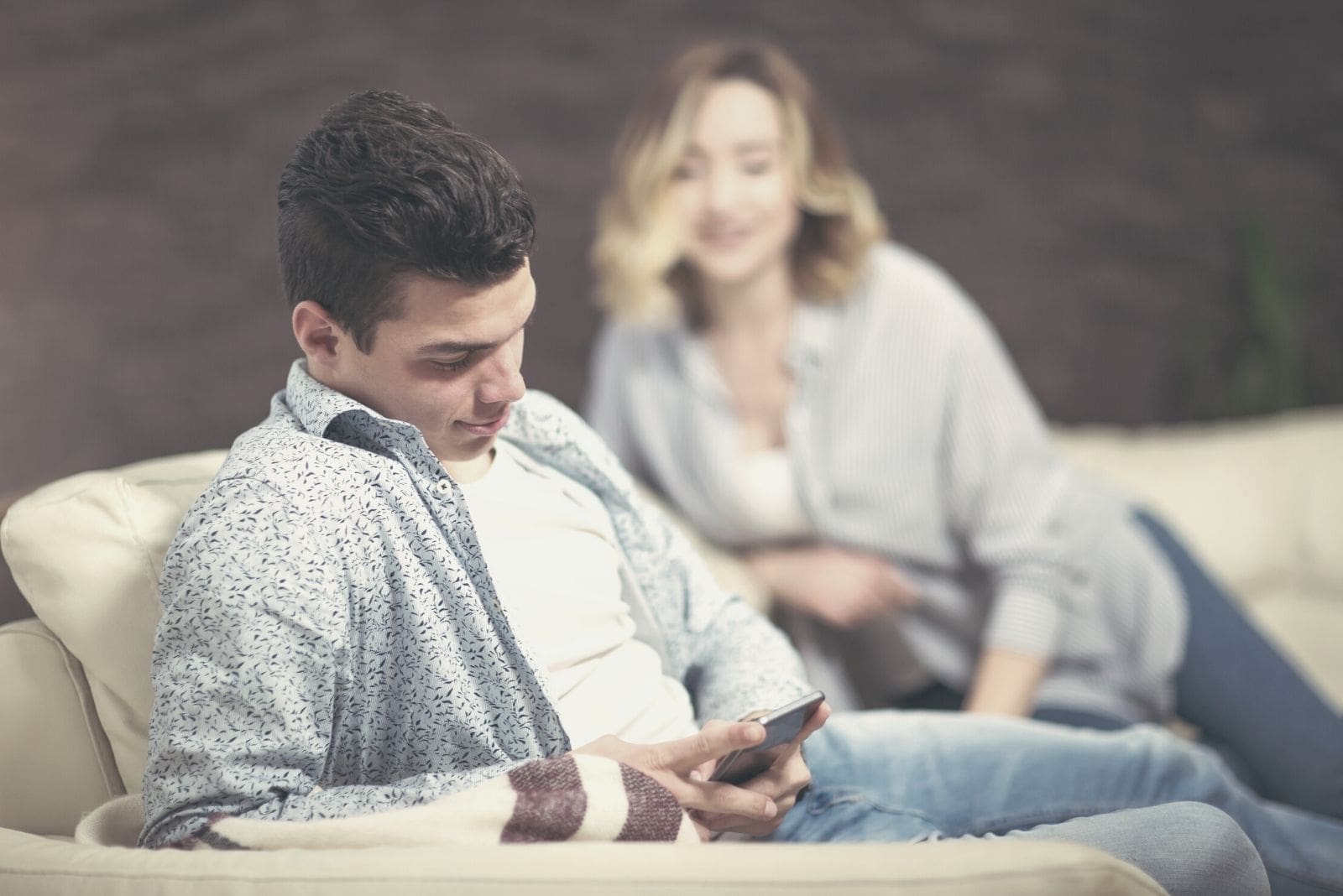 man texting in focus while sitting near a blurry woman snooping at his phone 