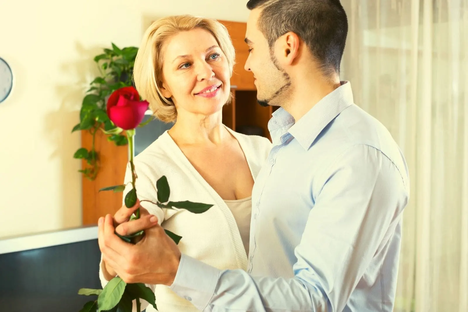 older woman dancing with a young man holding a rose inside the living room