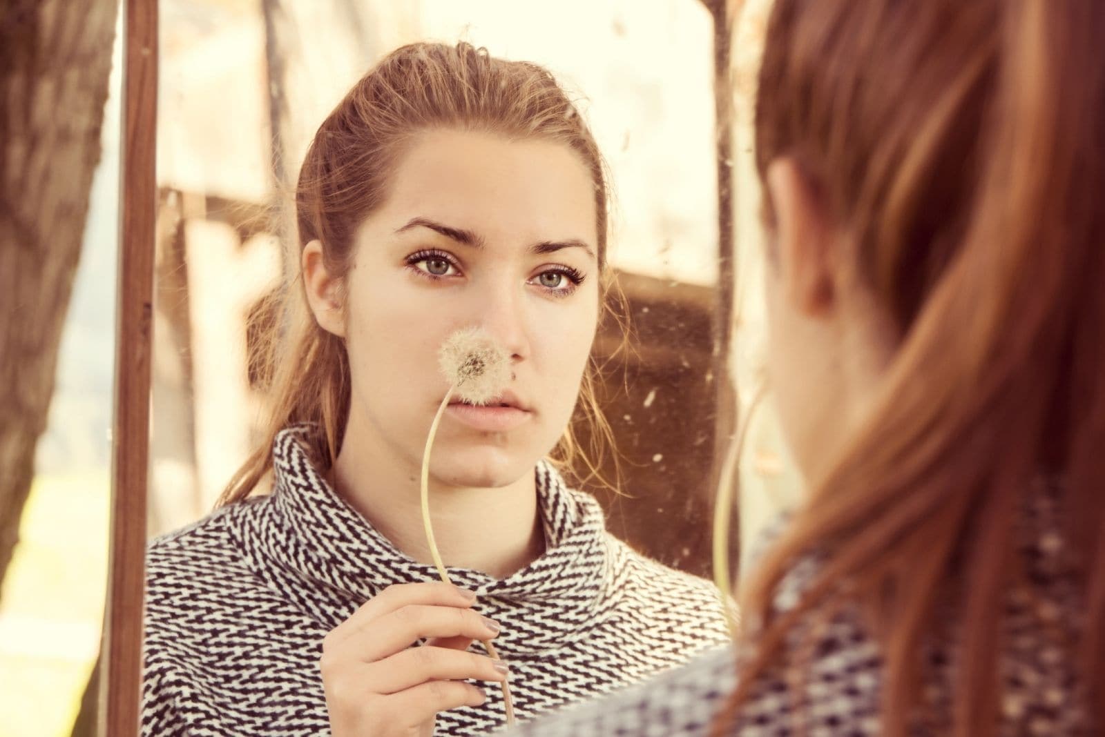 pensive woman holding a dandelion looking at herself at the mirror