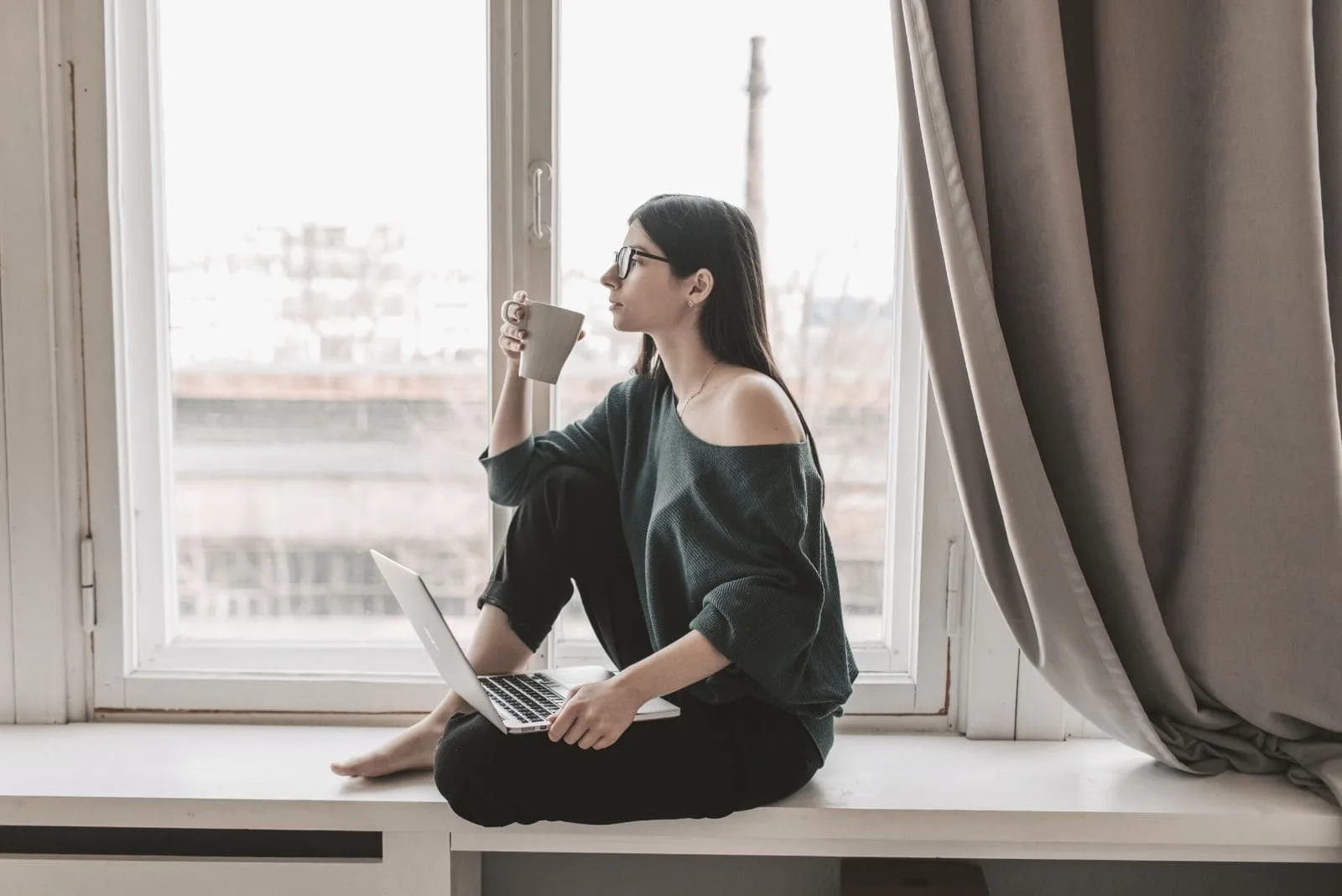 pensive woman with laptop drinking hot beverage sitting on the window sill