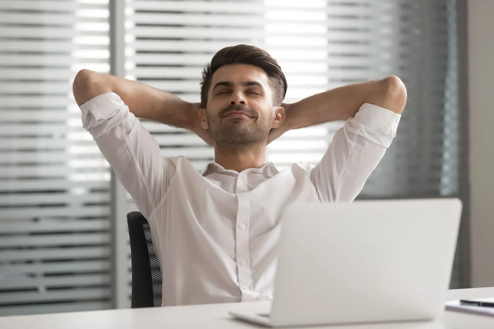 relaxed positive calm man sitting by the office table with both hands at the back of his head