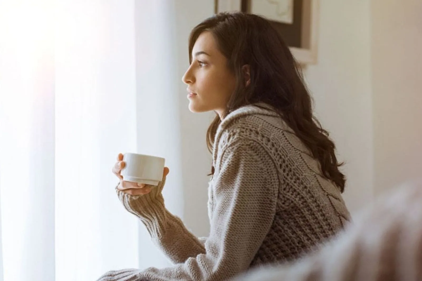 sideview of a young woman drinking coffee inside home looking out near the window pensively