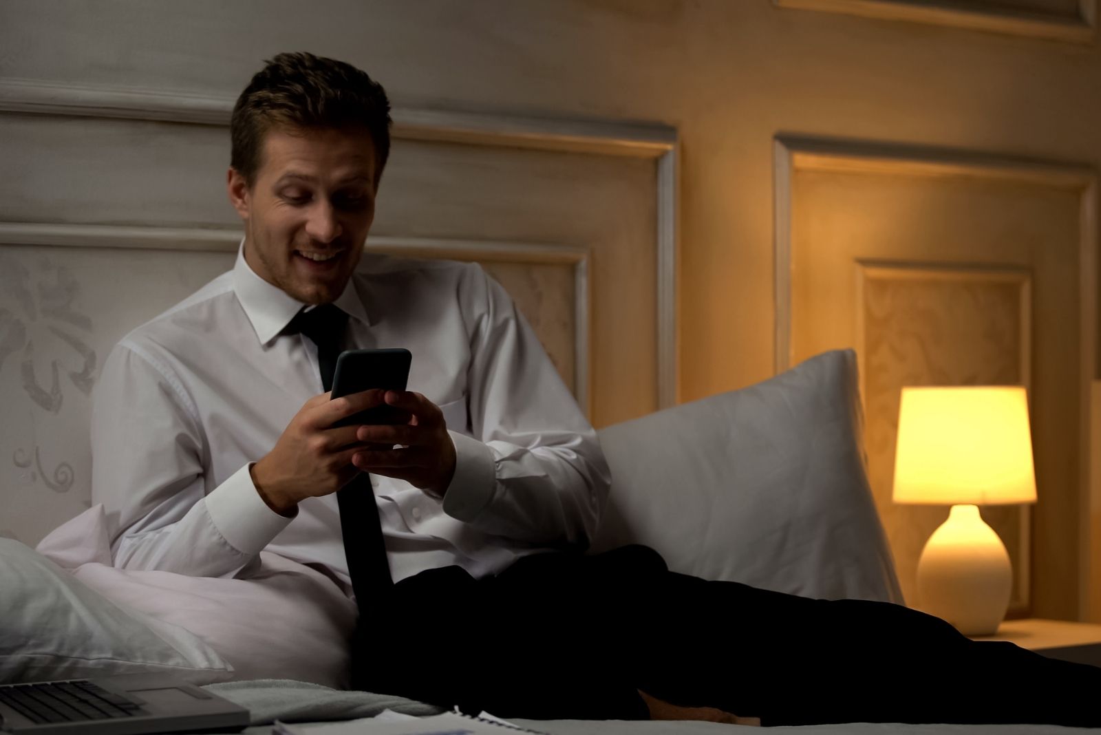 uomo sorridente in giacca e cravatta seduto sul letto con luci soffuse che messaggia con un'app di incontri