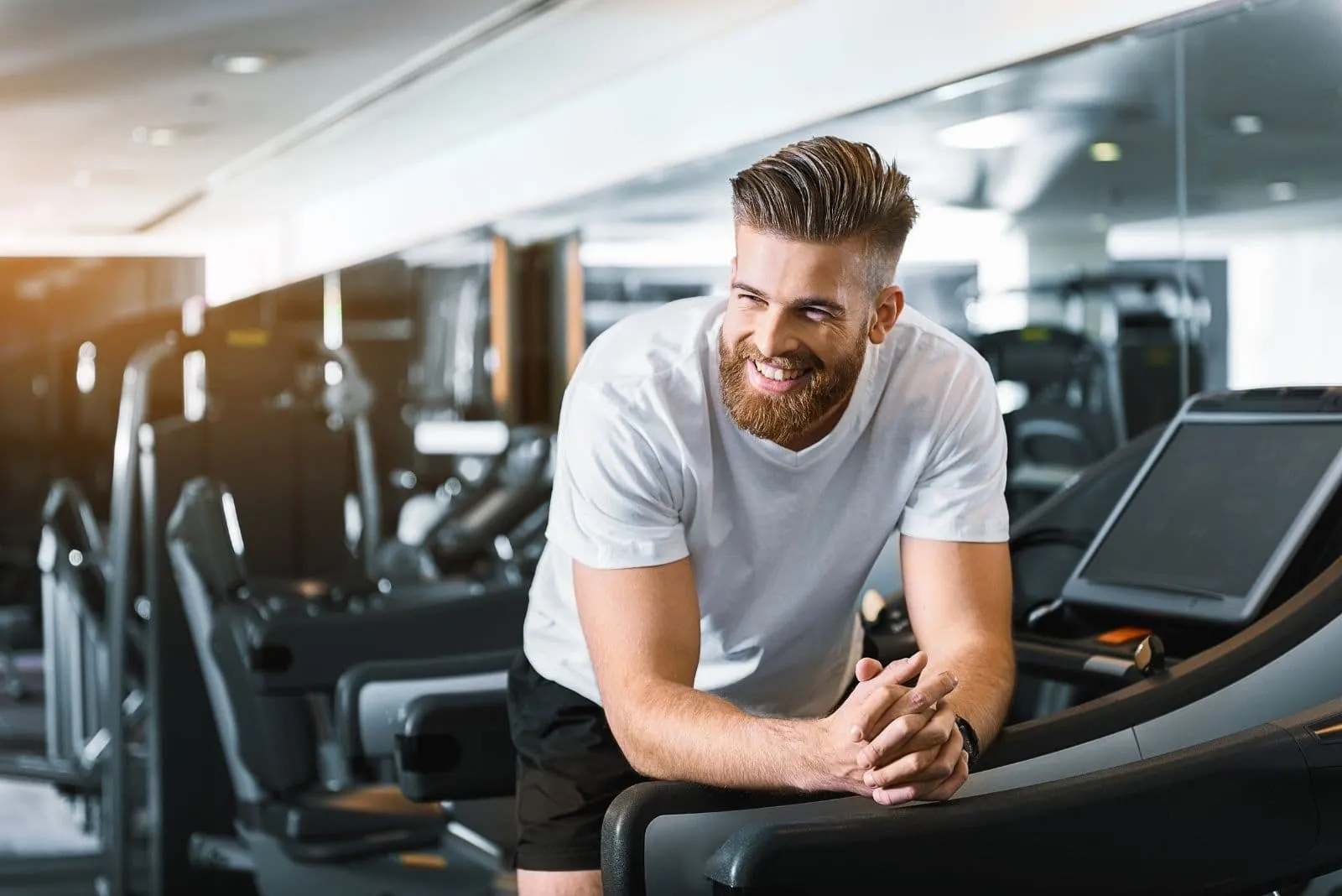 smiling man leaning on the side of the threadmill inside the gym