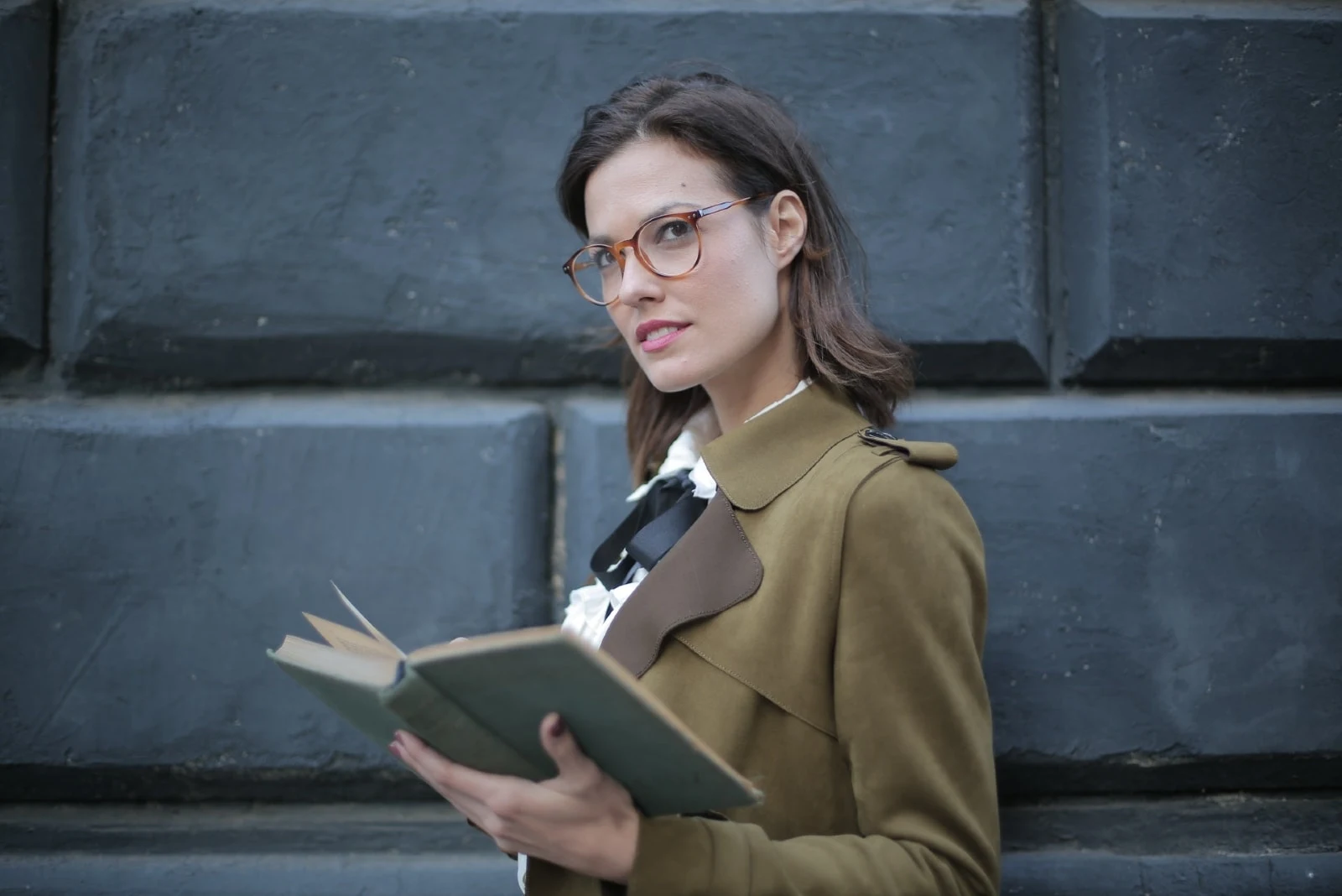 woman with eyeglasses holding open book while standing near wall