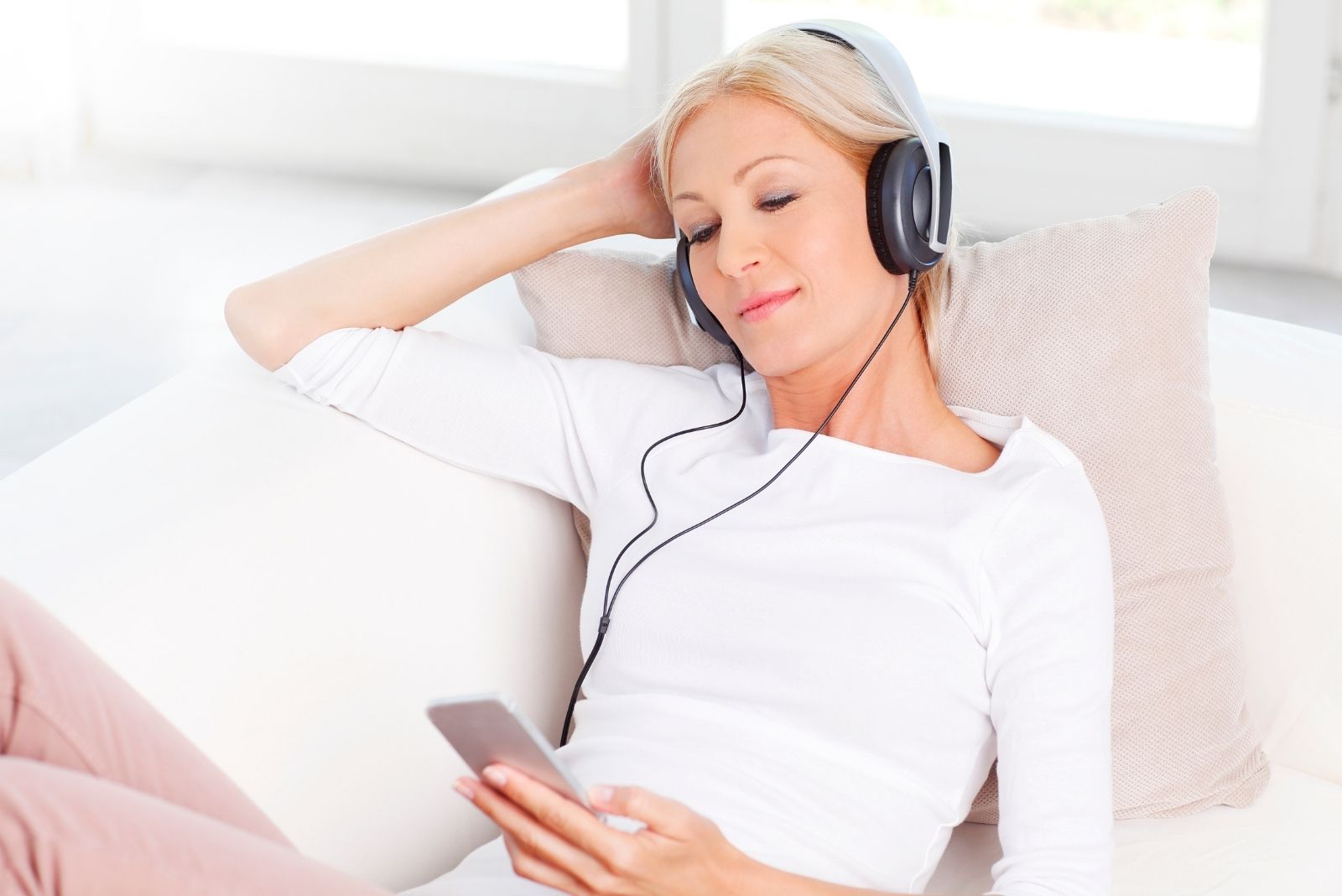 woman realxing holding a smartphone and listening with headphones on sitting on the couch near the windows