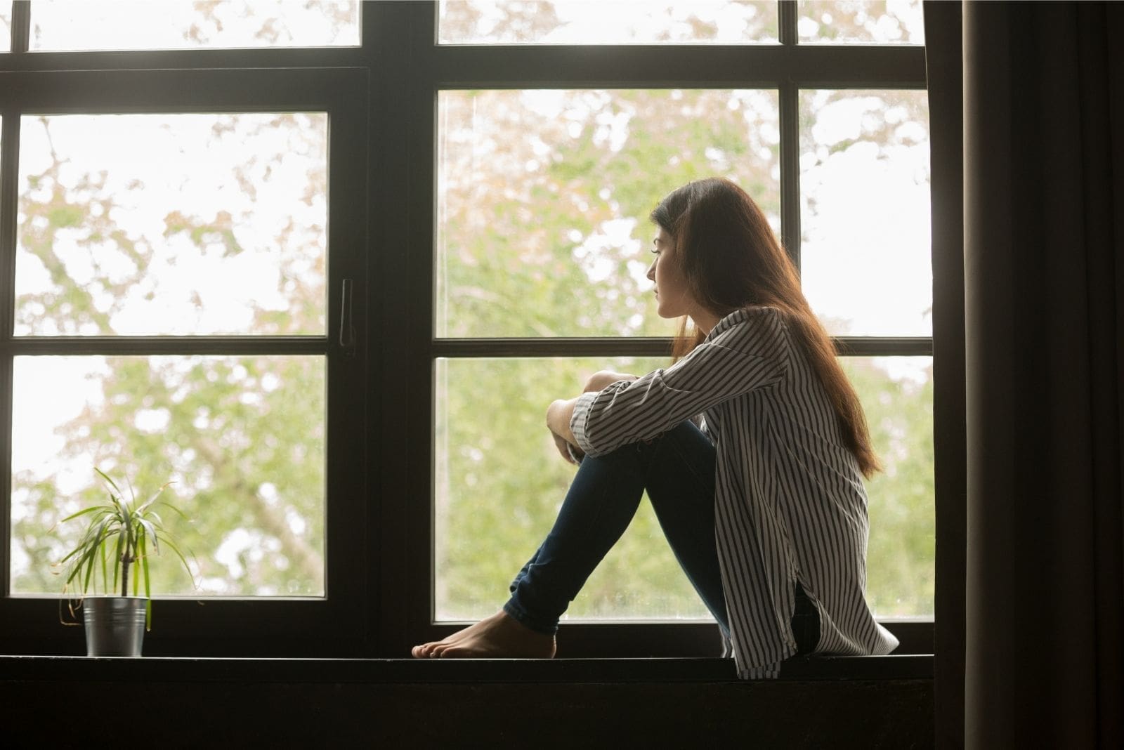 woman sitting by the window sill looking outsode the window wearing casual jeans and long sleeves