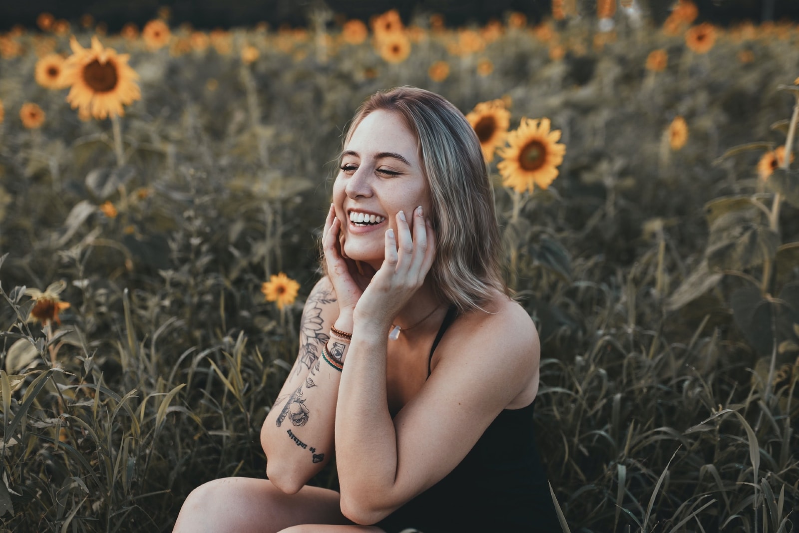 woman in black top laughing while sitting near sunflower field