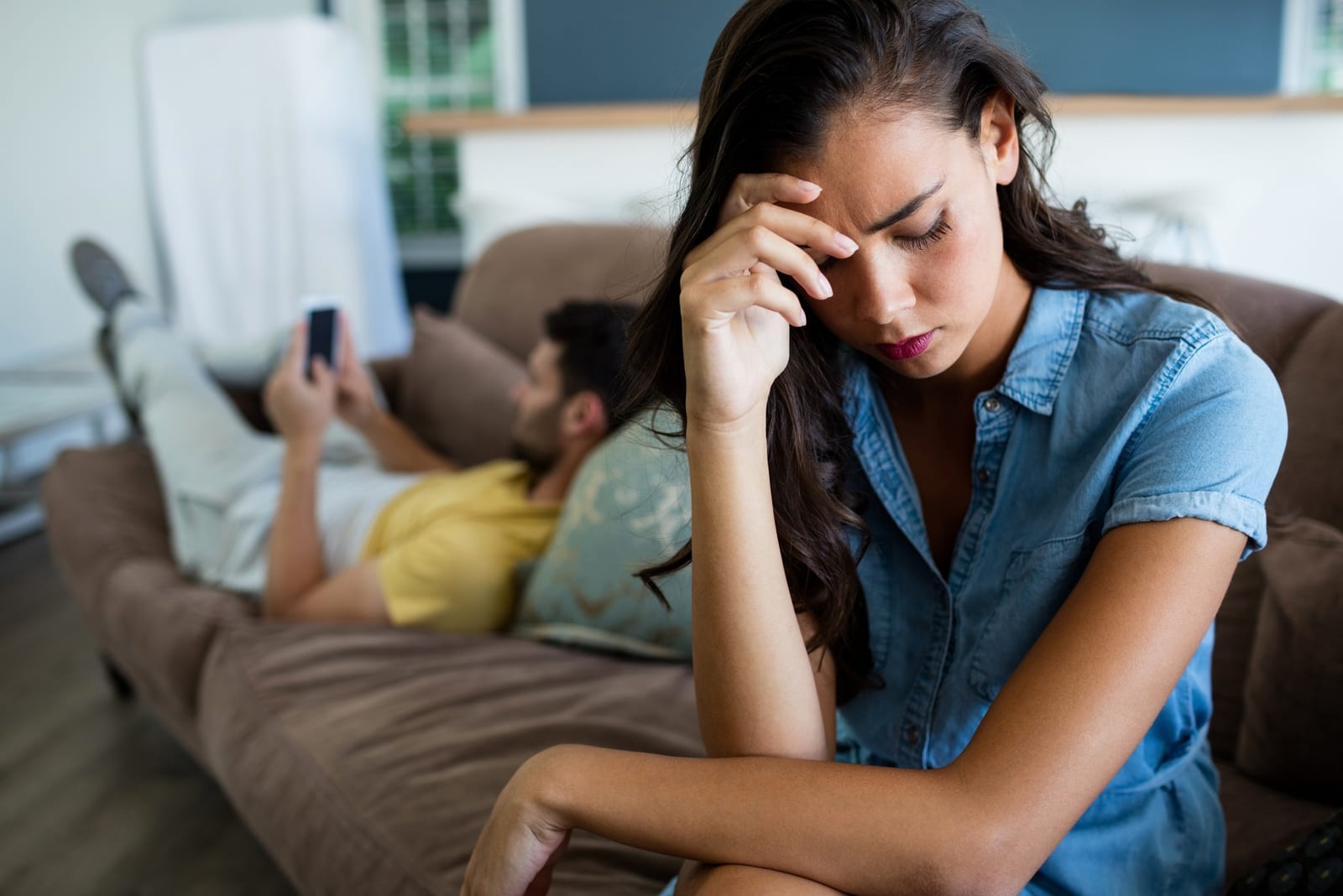 sad woman sitting on sofa while man using smartphone