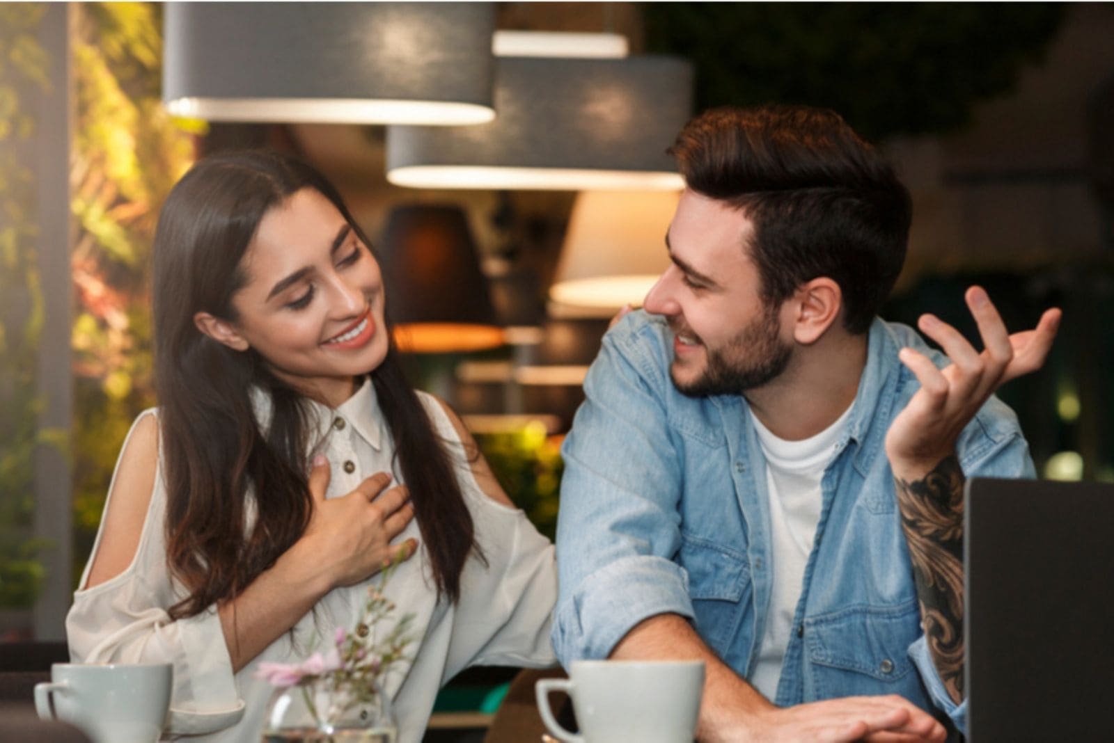 mulher a tocar no peito e a sorrir enquanto fala com um homem no café