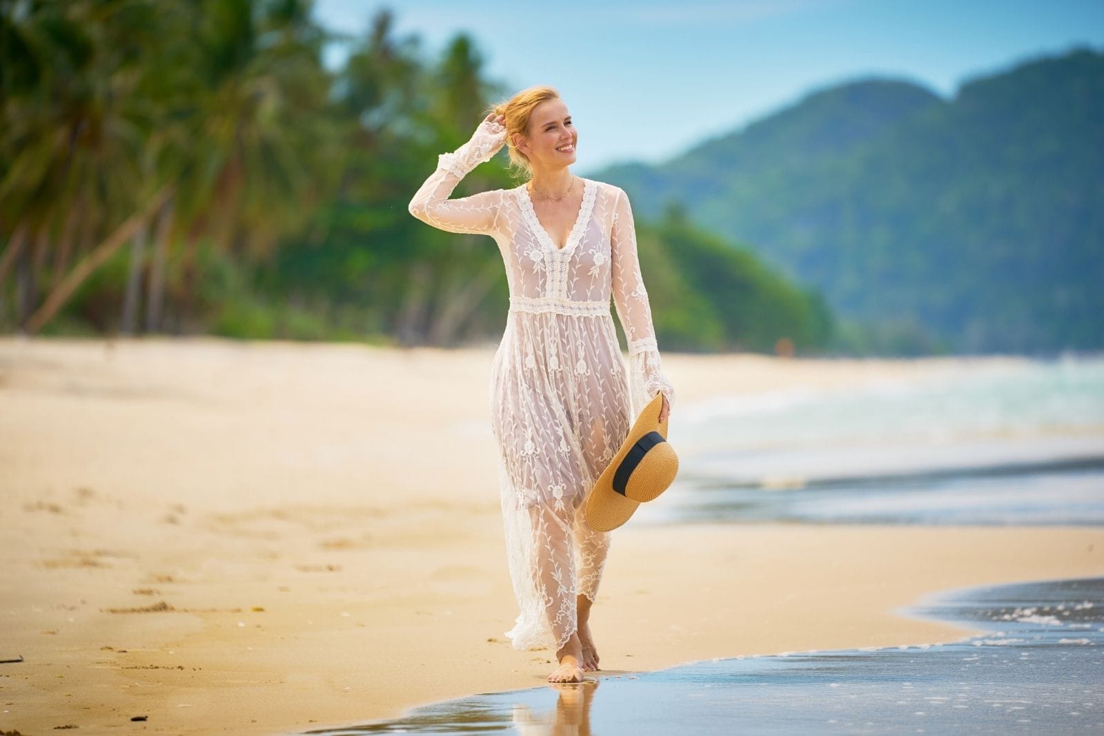 woman walking alone the shore of the beach trying to put her hair down and holding a hat smiling