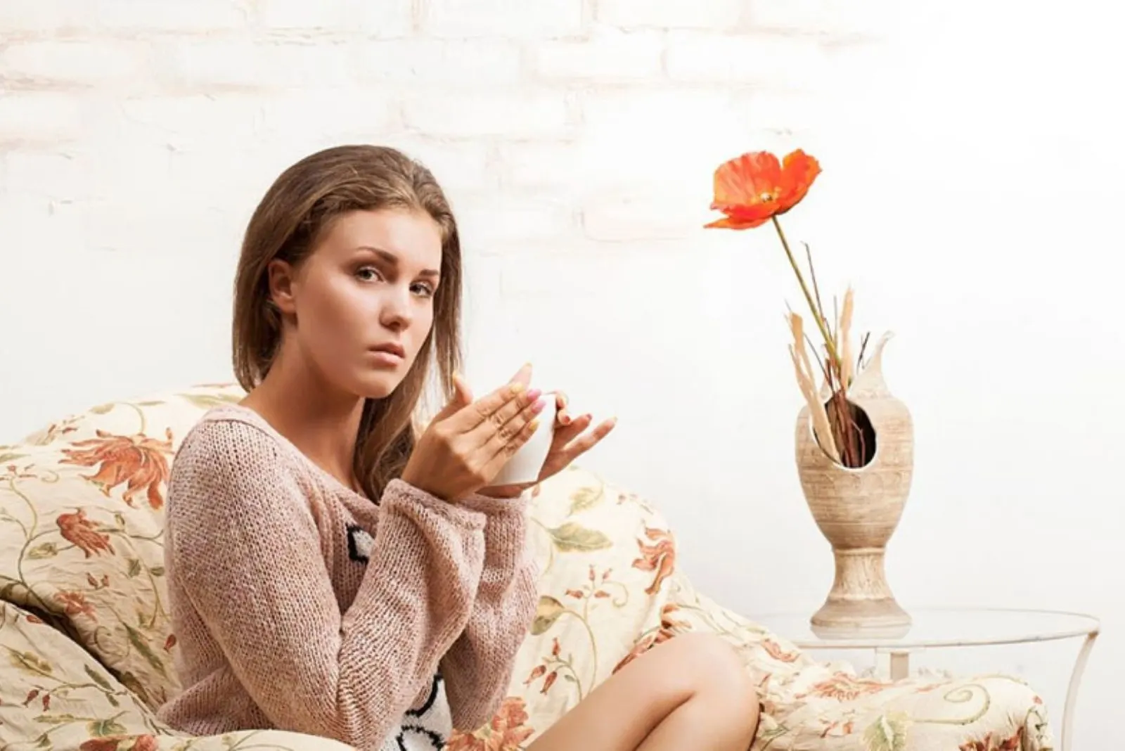 woman with a questioning eyes looking at the camera sitting on a couch and drinking a hot beverage