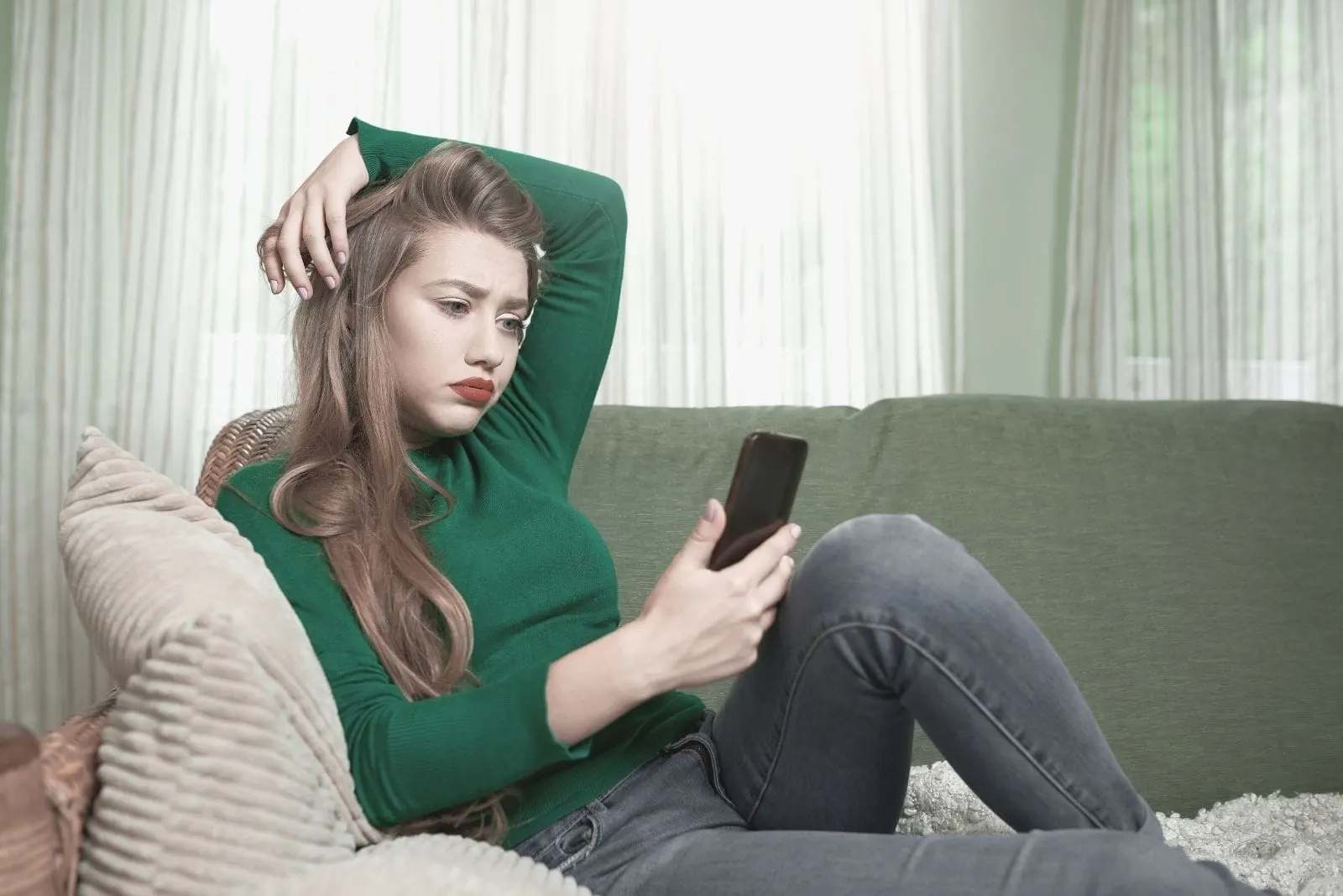 worried woman with her phone sitting on the couch putting her other arm on her head