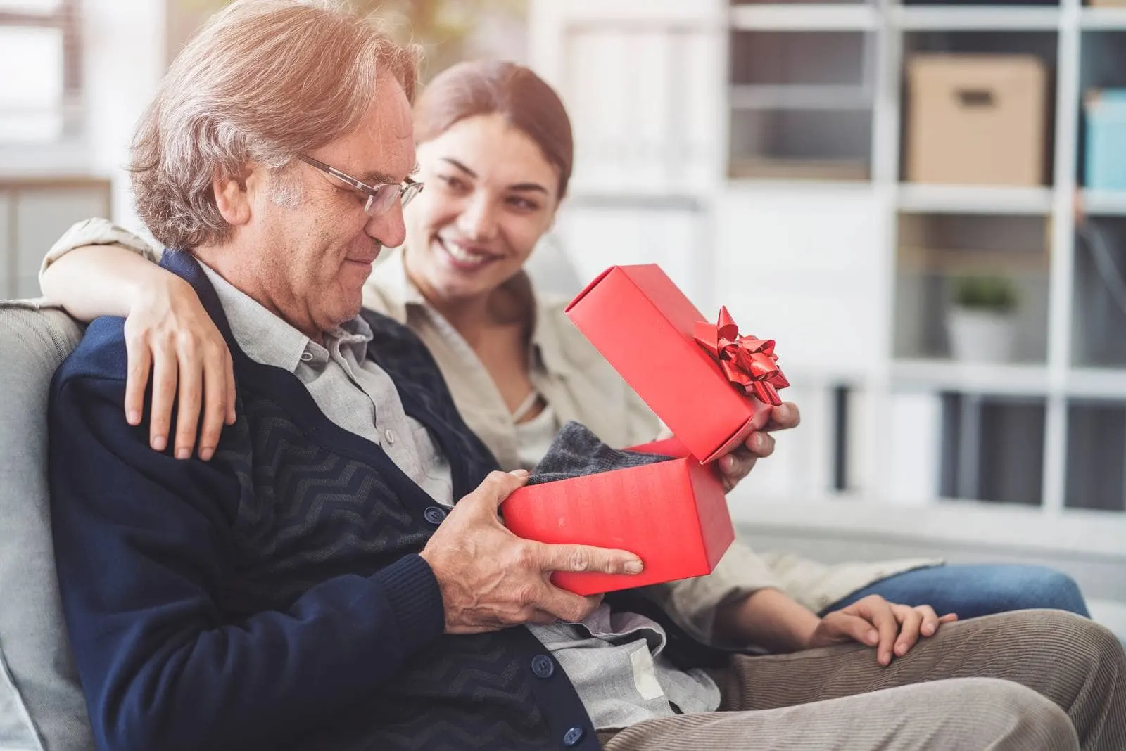 young loving daughter giving gift to her father sitting on the couch inside living room