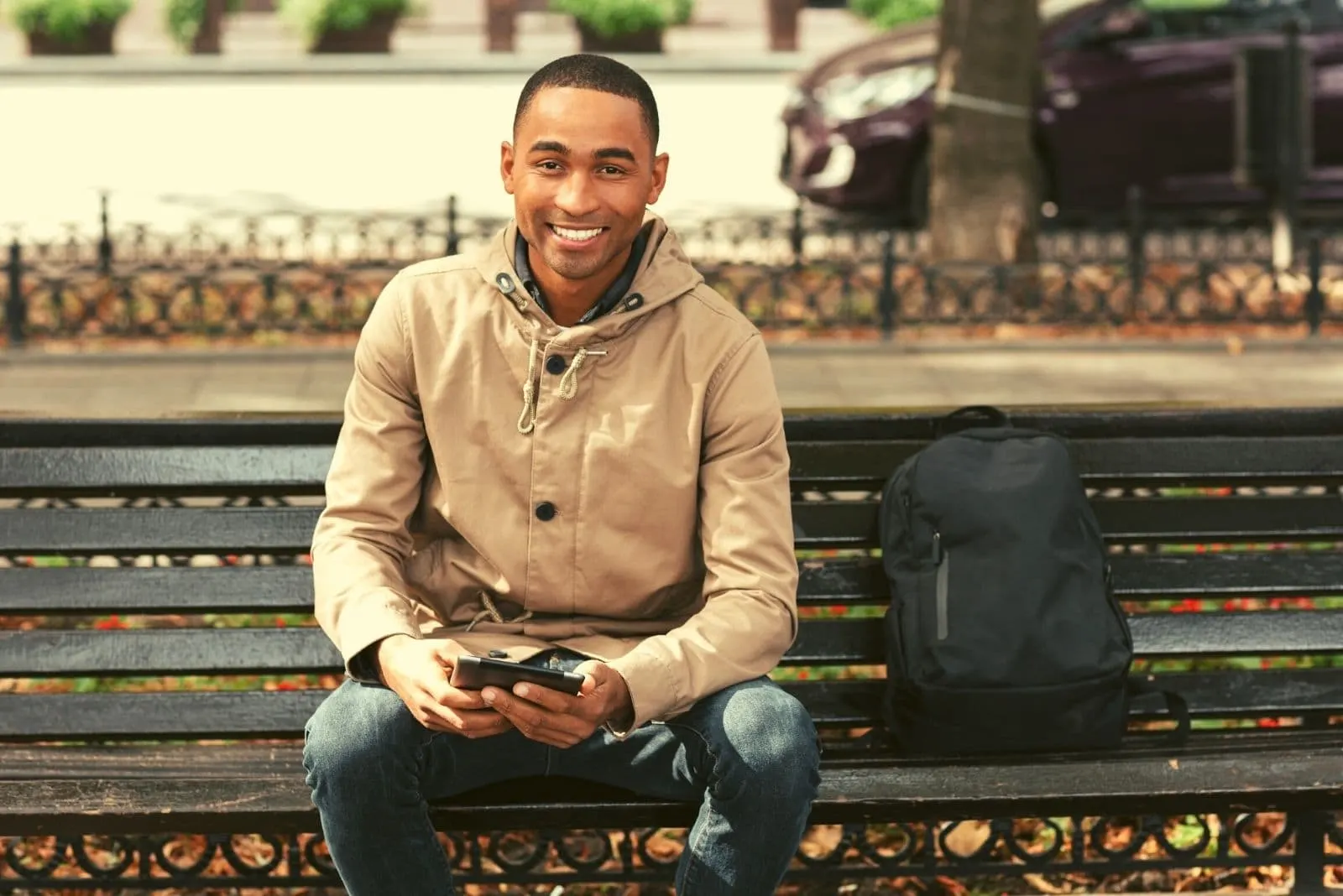 young smiling man using his tablet while sitting in the bench of the park and his bag beside him