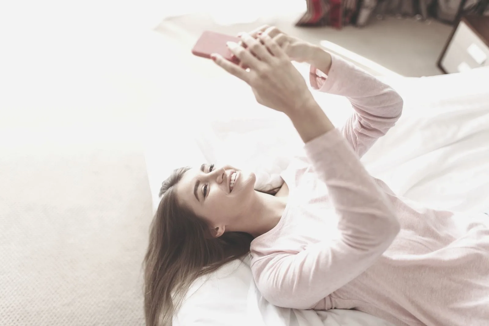 young woman lies in bed smiling and looking at her smartphone in the morning