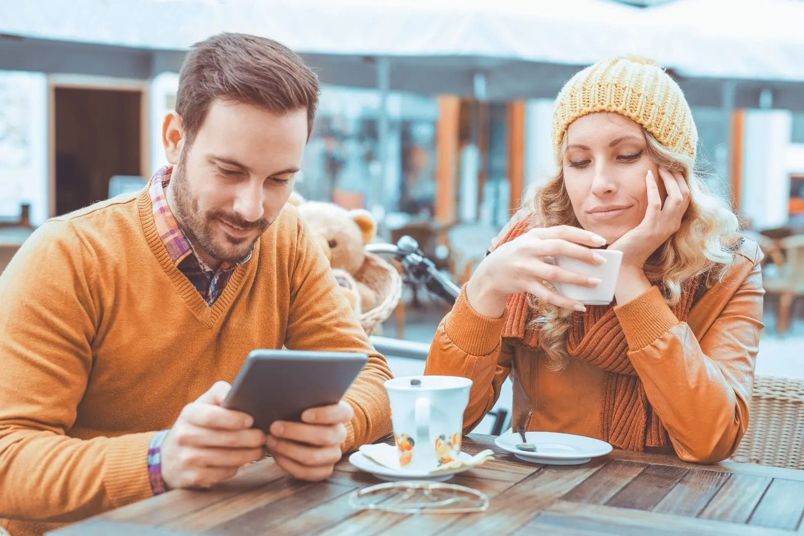 young woman looks suspicious while boyfriend is texting on smartphone while having coffee in an outdoor cafe