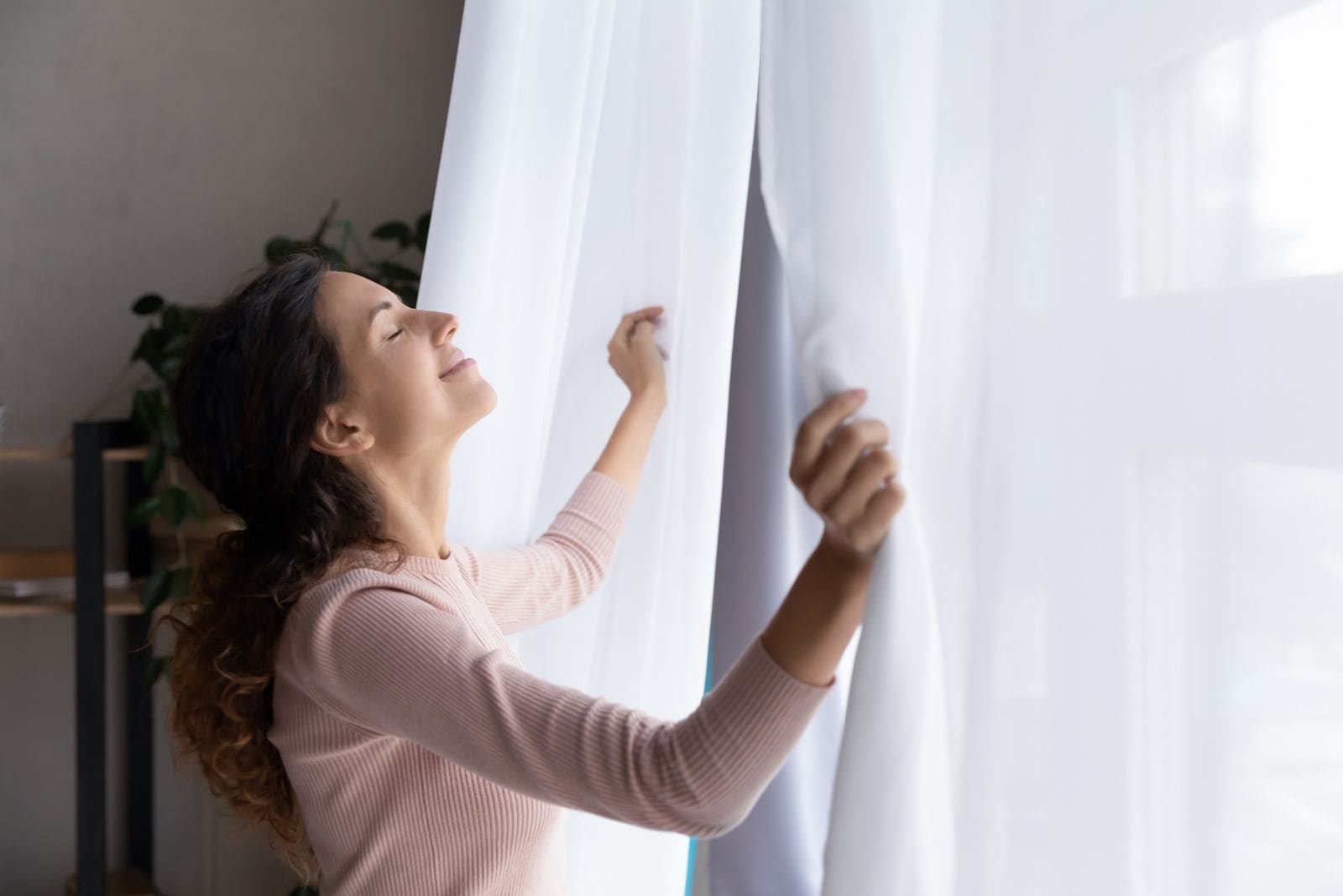 young woman opening curtains and closing eyes and smiling welcoming for a new day