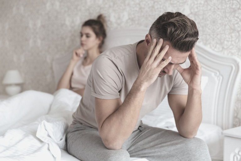 sad man sitting on bed with his girlfriend sitting in the background