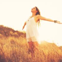 giovane donna felice e alla moda che sta in piedi e allarga le braccia in mezzo al campo durante il tramonto