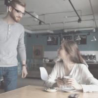 woman sitting in a cafe approached by a man with eyeglasses in her table