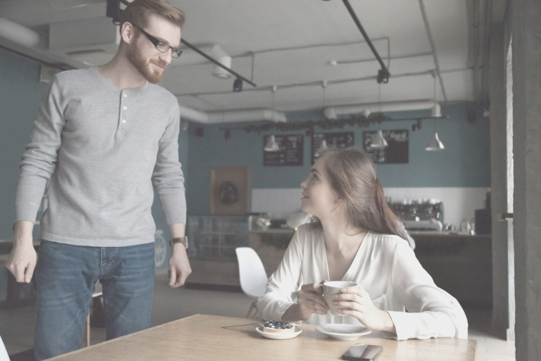 woman sitting in a cafe approached by a man with eyeglasses in her table
