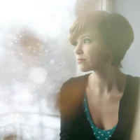 a sad short-haired brunette stands by the window and watches the snow fall outside