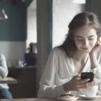 smiling woman reading messages from her smartphone inside the cafe with a blurry man at the back looking at her