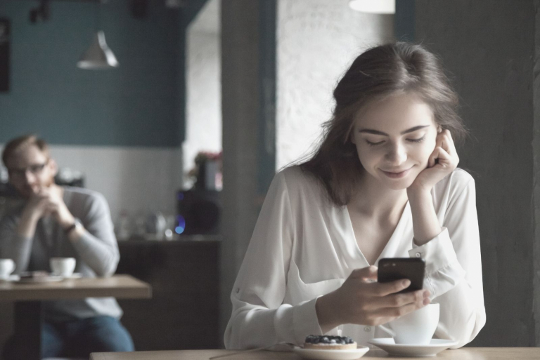 smiling woman reading messages from her smartphone inside the cafe with a blurry man at the back looking at her