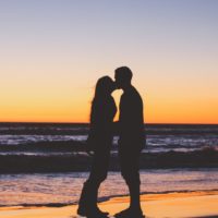 man and woman kissing while standing on beach during sunset