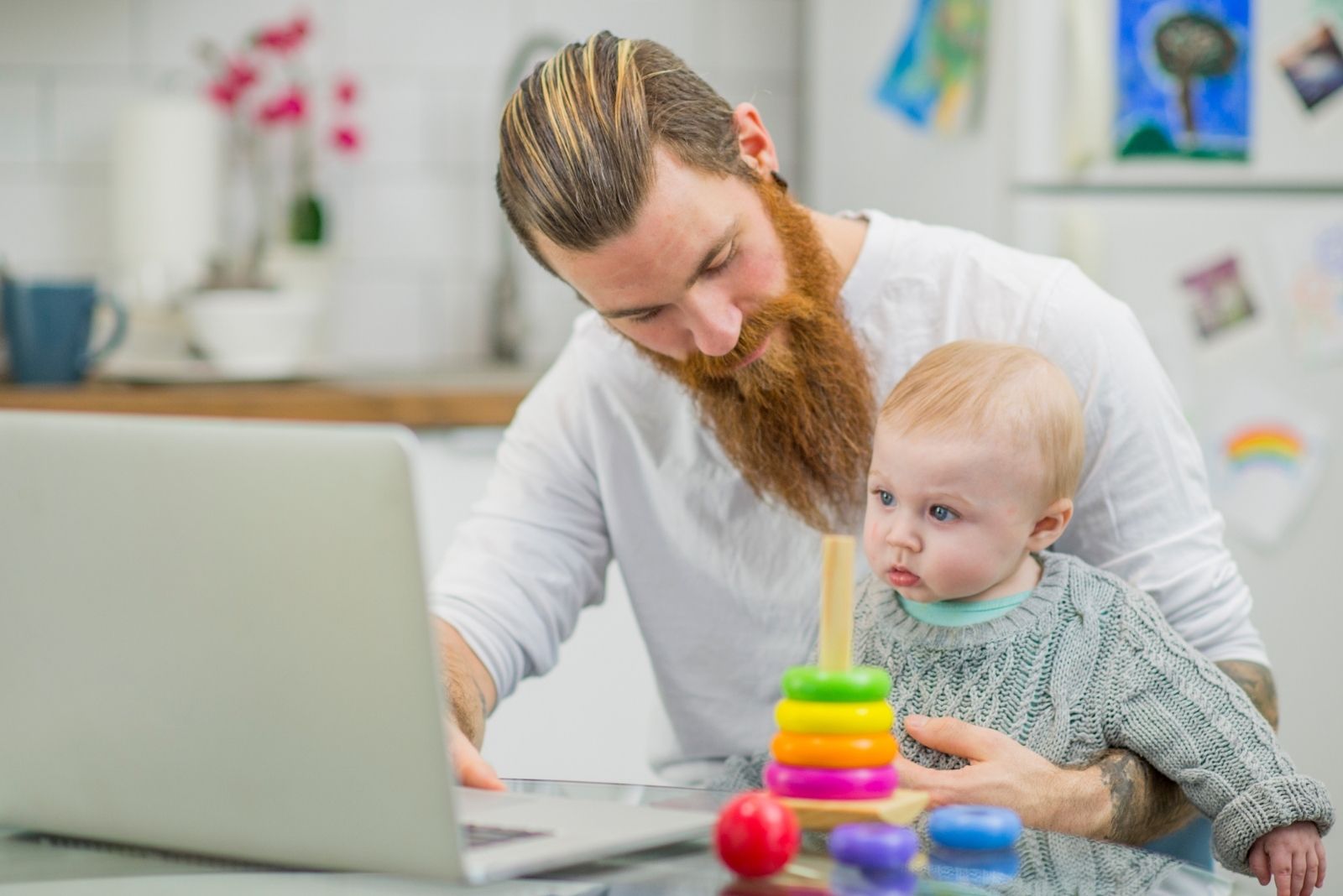 un giovane padre caucasico che lavora al suo laptop con un bambino in grembo e dei giocattoli sul tavolo