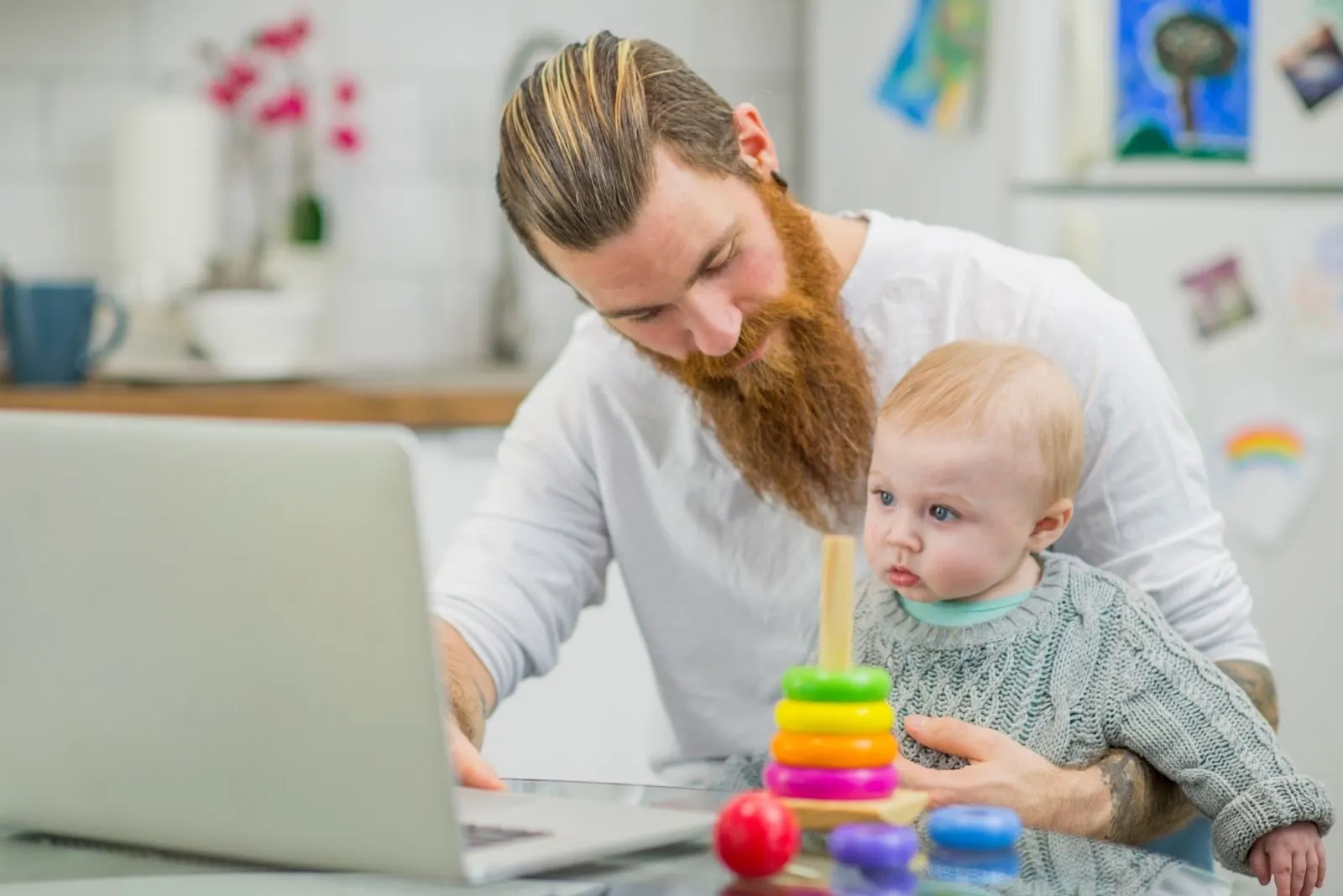 a young caucasian father working on his laptop with a baby on his lap and toys on the table