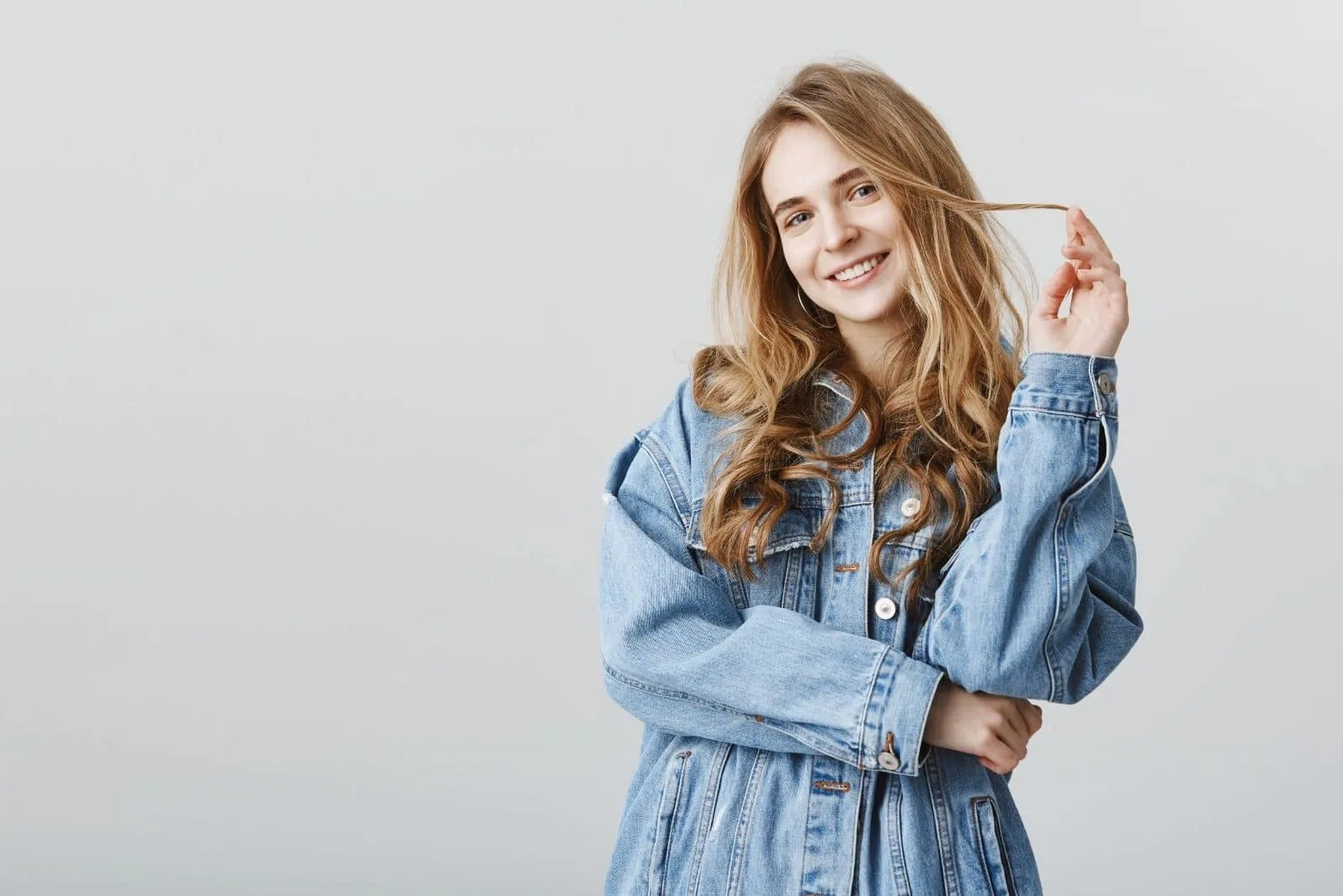 beautiful caucasian woman standing wearing oversized denim jacket and playing with her hair and smiling