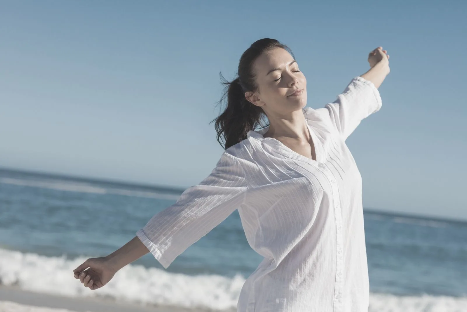 calm woman stretching in beach wearing white long sleeves and closing her eyes