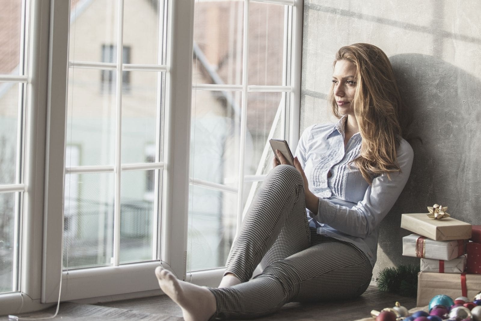 caucasian woman is sitting on the floor near the big glass windows holding a phone