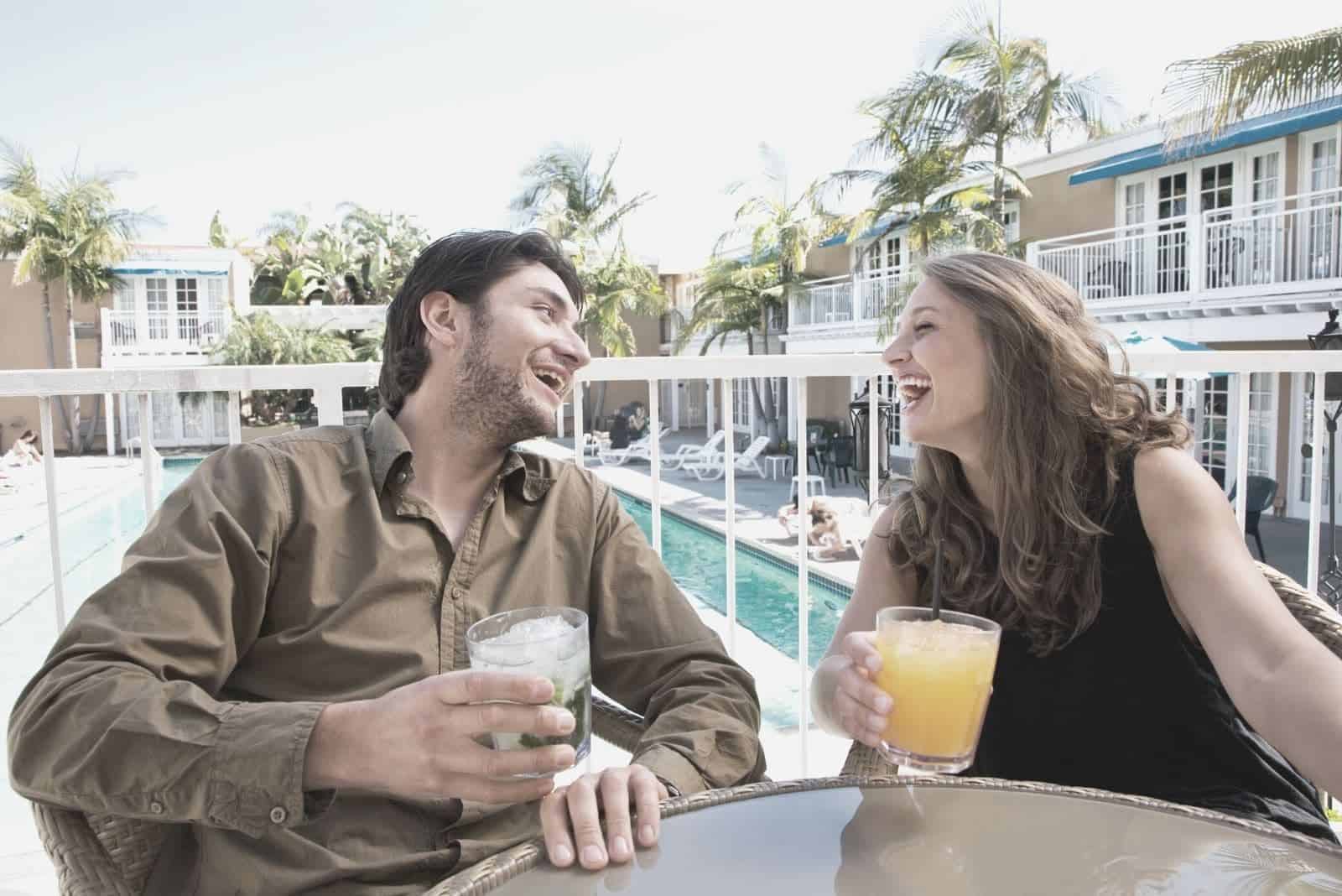 alegre pareja riendo mientras toma una copa y se sienta junto a la mesa en un porche cerca de una piscina