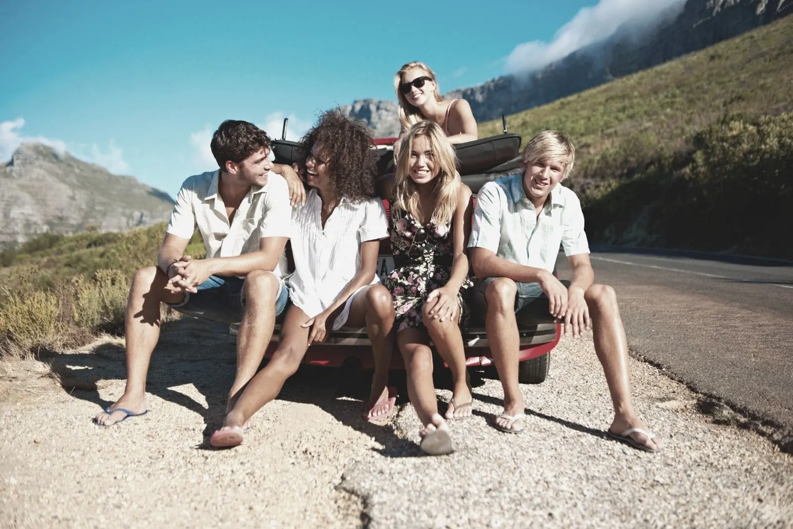 group of young friends sitting on the car bump while on a road trip