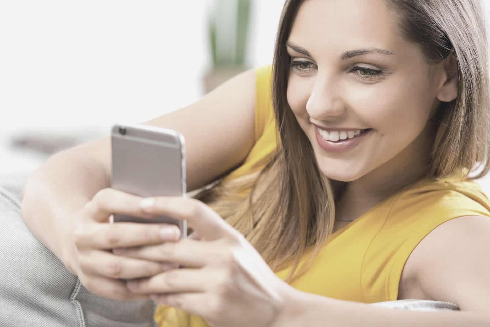 mujer feliz enviando mensajes de texto y sonriendo mientras descansa en un sofá