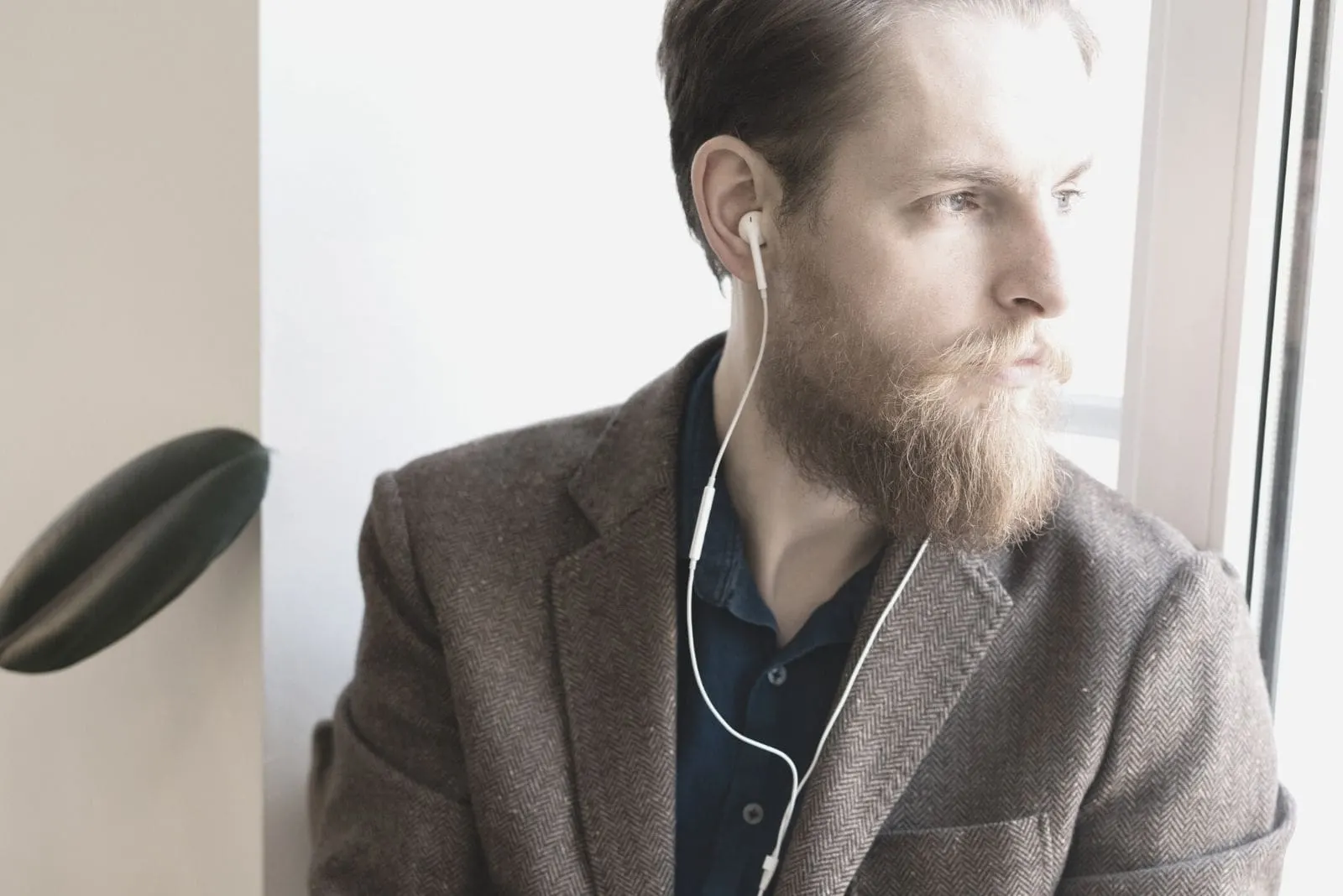 introspective hipster man with beard sitting in the window sill using a earphone