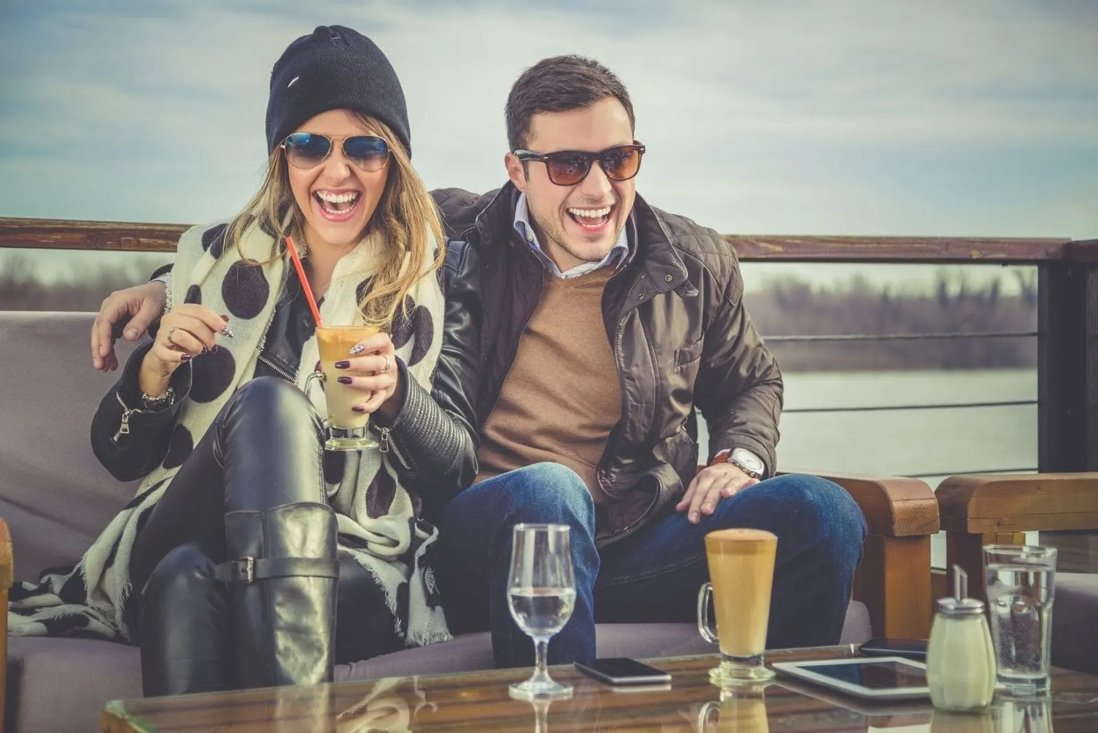 man and woman laughing wearing winter clothes sitting near the body of water and drinking beverage