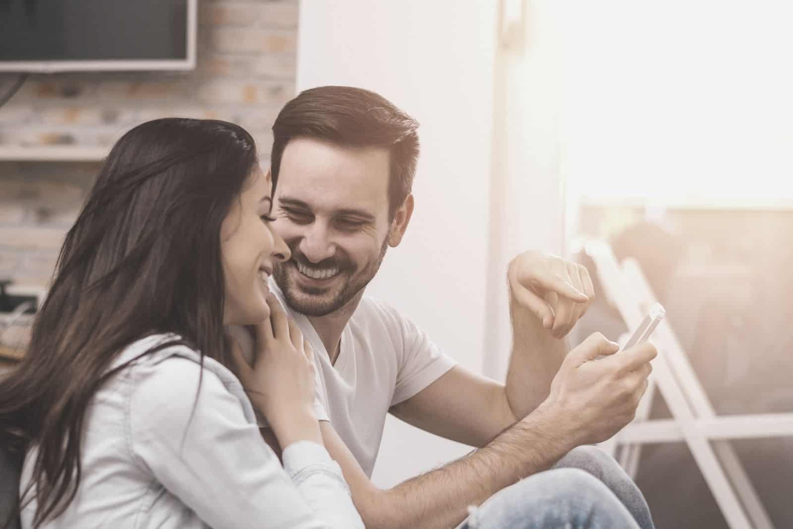 homem com um smartphone na mão mostrando-o a uma mulher que se inclina e ri com ele, sentado na varanda